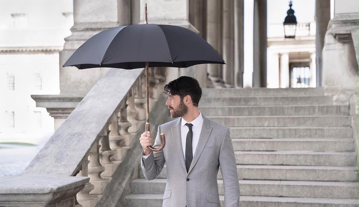 A man in a grey suit holding a black Bucklesbury London Umbrella with a curved wooden handle on stone steps in London