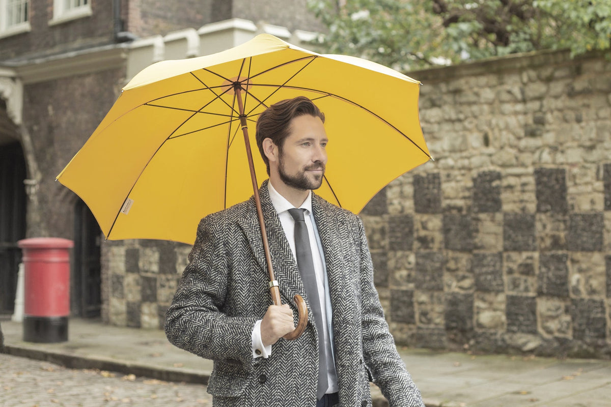 A man in a grey tweed coat holding an open yellow Bucklesbury Classic Umbrella with a curved wooden handle on a London street