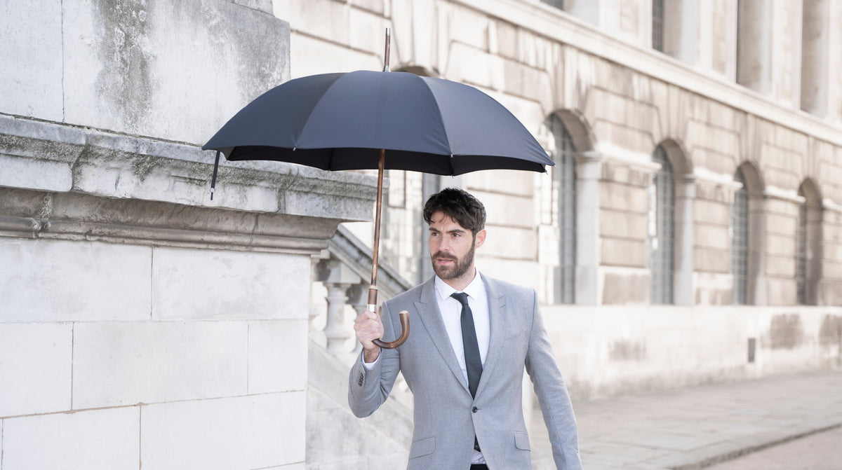 A man in a grey suit walking past white stone buildings in London under a black Bucklesbury London Umbrella with a wooden crook handle